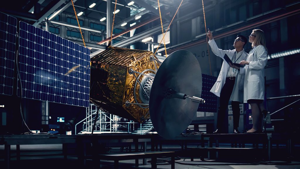 Zwei Wissenschaftler in Laborkitteln untersuchen und diskutieren einen Satelliten in einer großen Industrieanlage, umgeben von heller Beleuchtung und Geräten, auf denen Sonnenkollektoren und eine große Schüssel zu sehen sind.