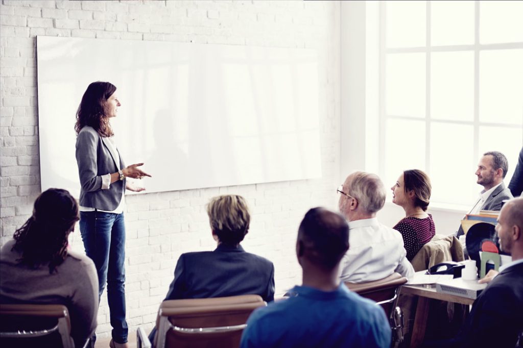 Eine Frau steht und spricht vor einer Gruppe von sitzenden Erwachsenen in einem Büro oder Klassenzimmer, mit einer leeren Tafel hinter ihr und großen Fenstern, die natürliches Licht hereinlassen.