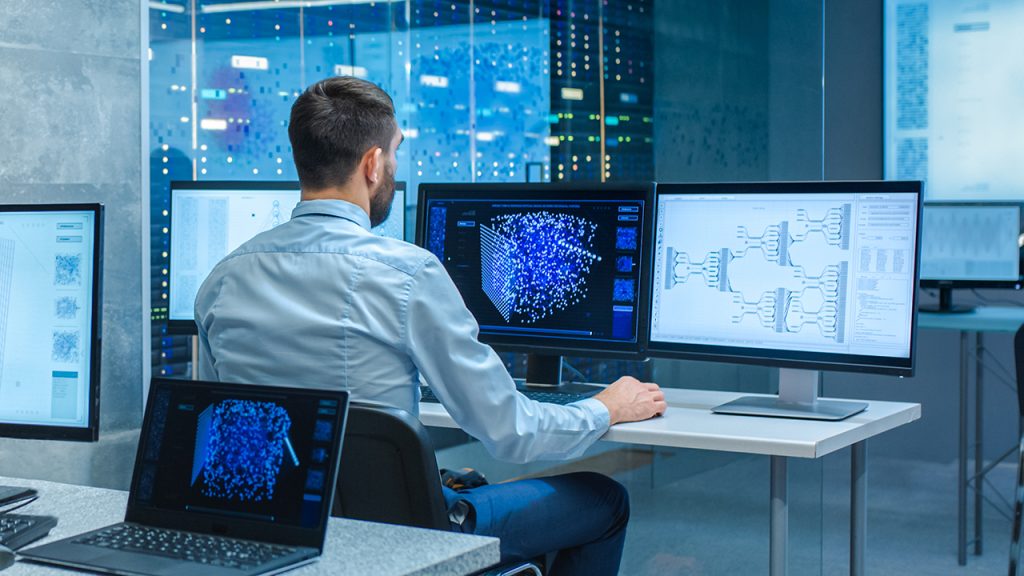 Ein Mann sitzt an einem Schreibtisch in einem Serverraum und analysiert Datenvisualisierungen und Netzwerkdiagramme, die auf mehreren Computermonitoren und einem Laptop angezeigt werden.