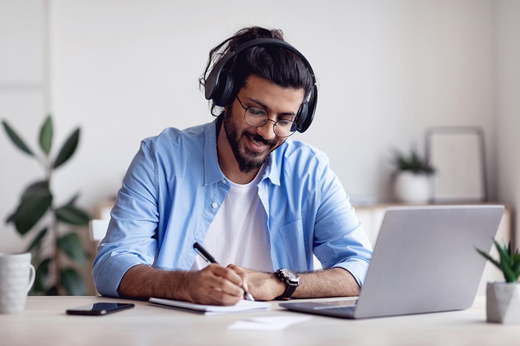 A man wearing headphones and glasses smiles while writing in a notebook at a desk with a laptop. He is casually dressed in a blue shirt, sitting in a bright room with plants and décor in the background.