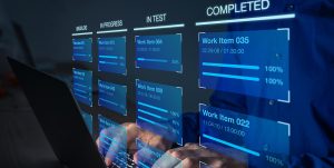 Person typing on a laptop with a virtual project management dashboard displayed, showing columns labelled "Backlog," "In Progress," "In Test," and "Completed" with work items and progress bars.