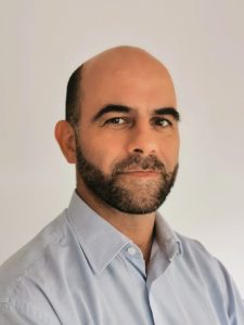 A man with a neatly trimmed beard and moustache, bald on top, wearing a light blue collared shirt, poses against a plain, light-coloured background, looking slightly to the side and smiling gently—inviting you to contact us.