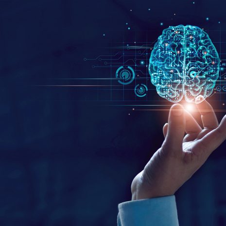 A hand reaching up with fingers touching a glowing digital brain hologramme, surrounded by circuit patterns and icons, representing artificial intelligence and technology on a dark background.